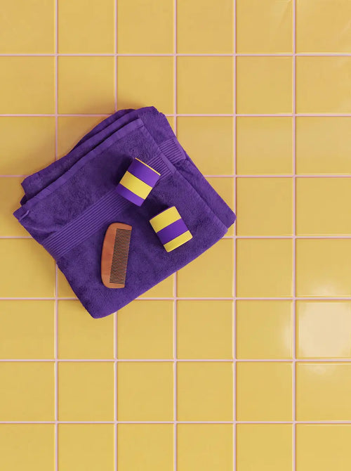 Two silicone travel cases in purple and yellow on a folded purple towel with a wooden comb