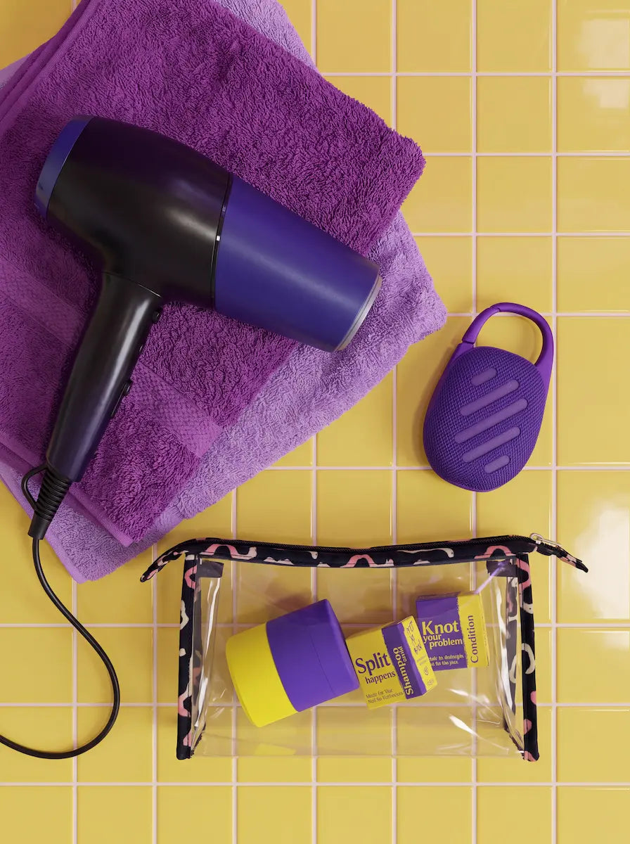 Silicone travel case and travel-size bars packed in a clear toiletry bag next to a hair dryer and towel