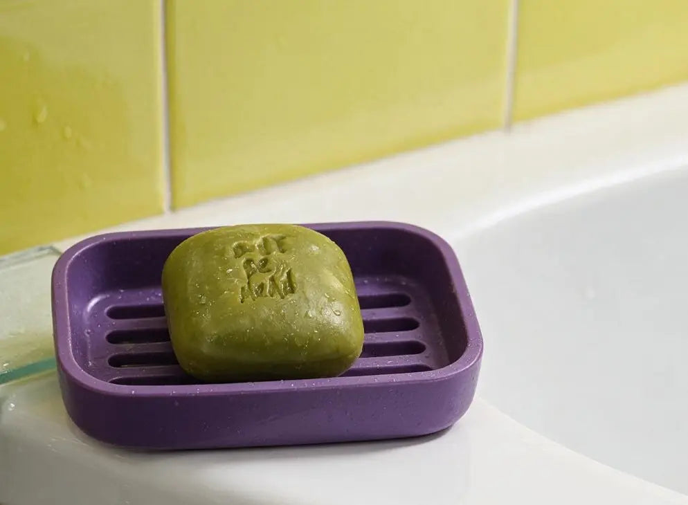 Green solid shampoo bar in a purple soap dish on a bathroom sink