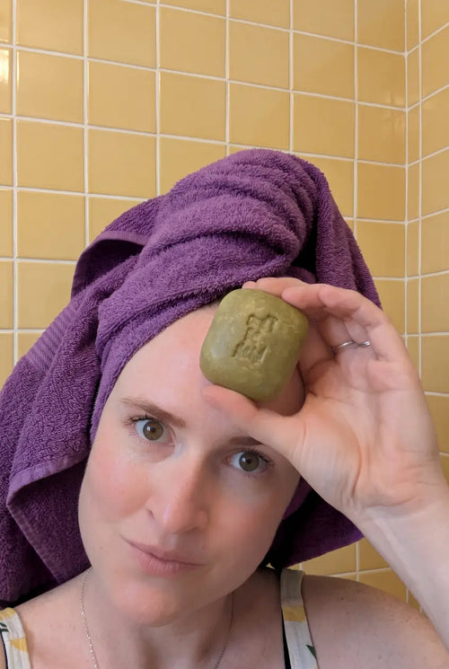 Woman with a purple towel on her head holding a green solid shampoo bar