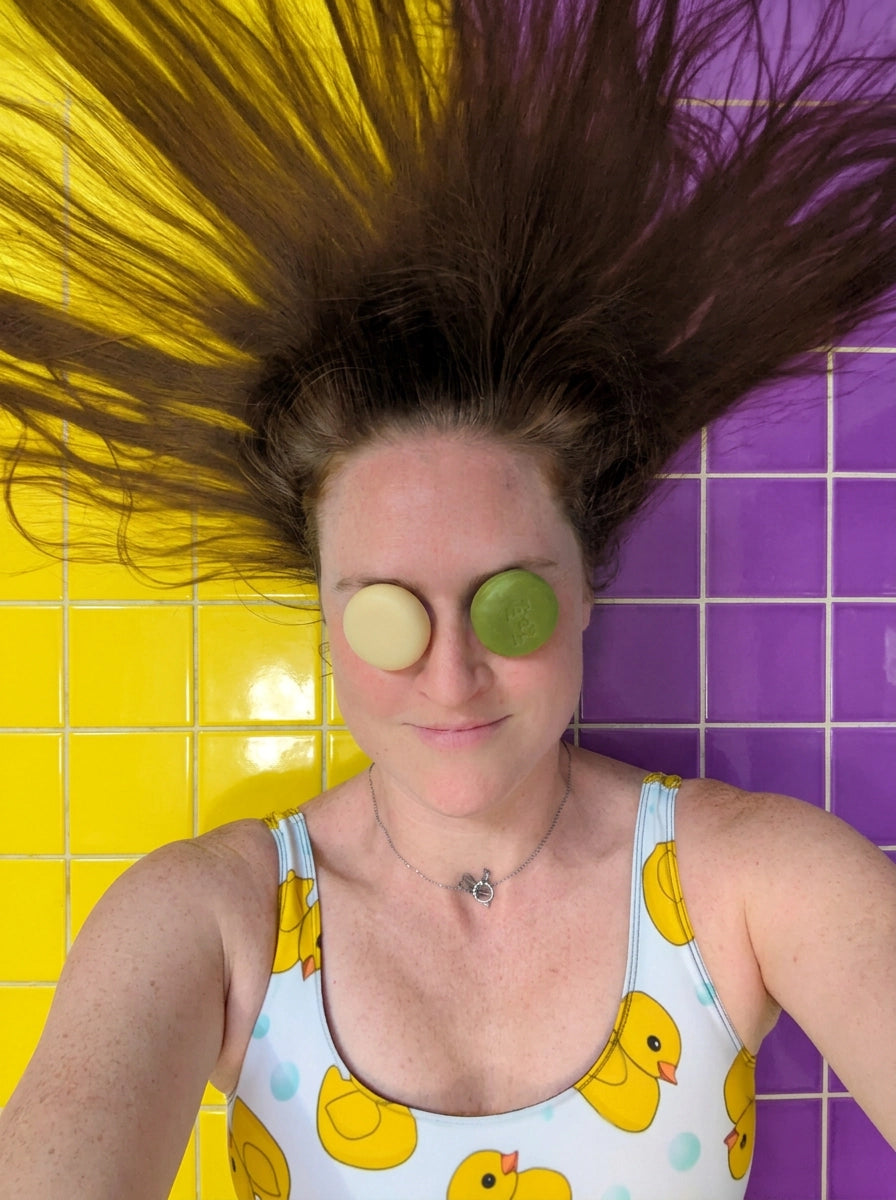 Woman with hair spread out on yellow and purple tiles holding travel-size shampoo and conditioner bars over her eyes