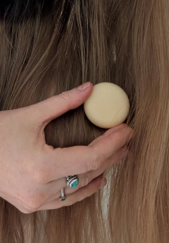 Hand holding a round travel-size conditioner bar against long brown hair