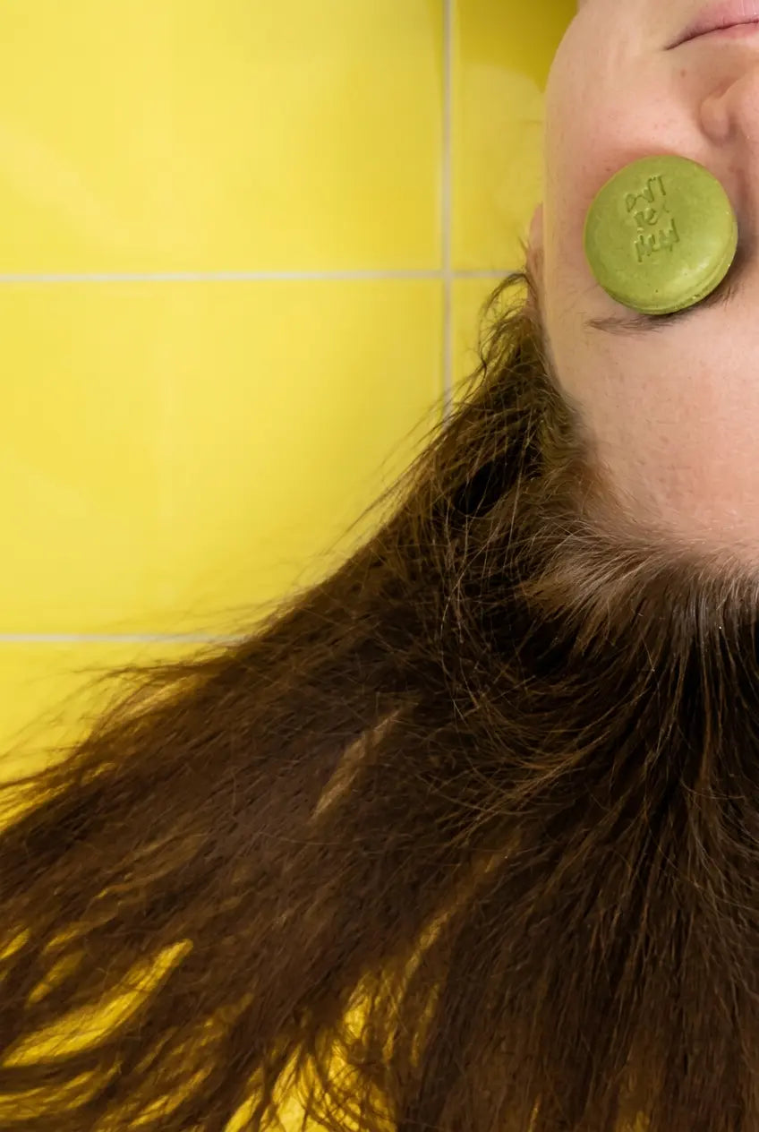 Round green travel-size shampoo bar held over an eye, long hair on yellow tiles