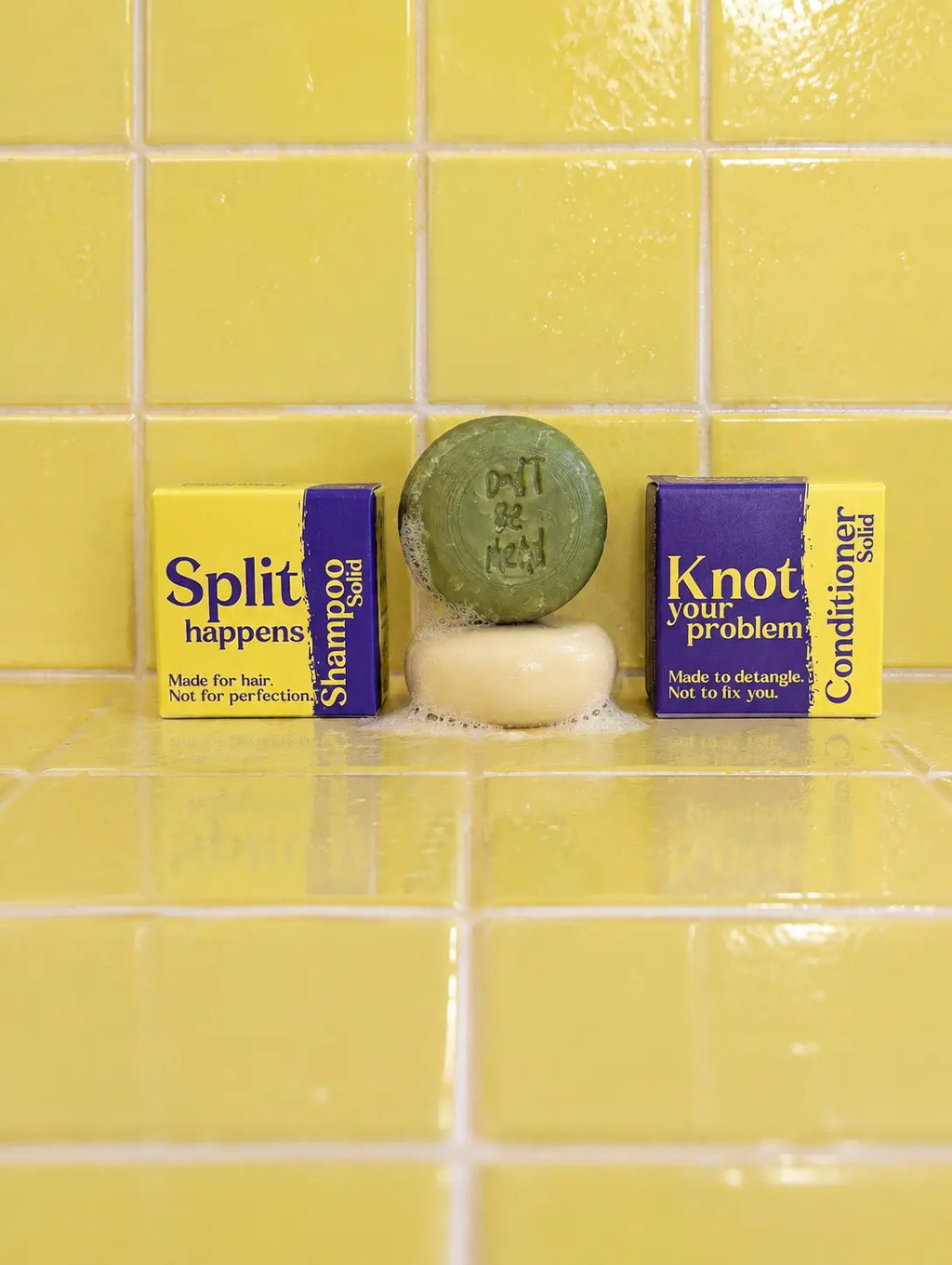 Travel-size solid shampoo and conditioner bars stacked between their boxes on yellow tiles