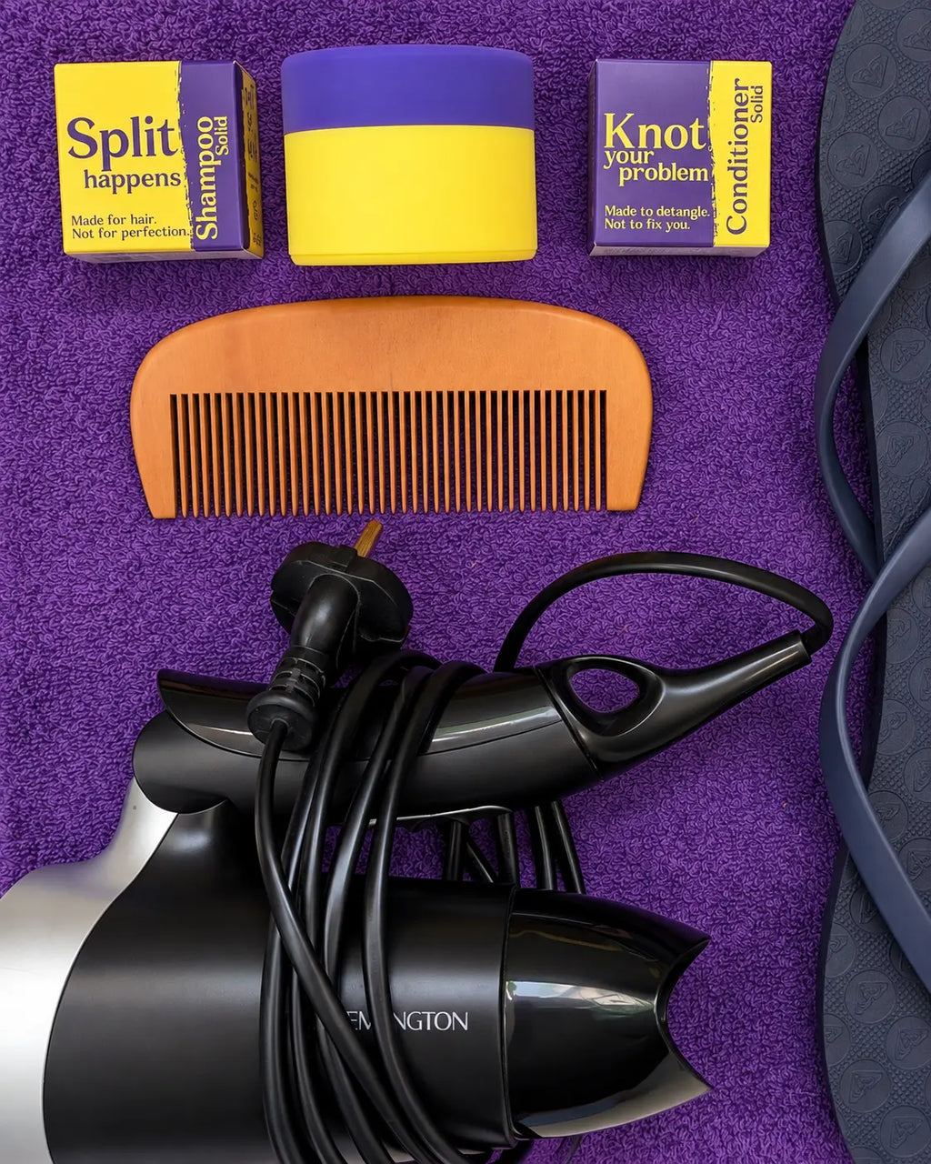Travel shampoo and conditioner boxes with a silicone case, comb and hair dryer on a purple towel