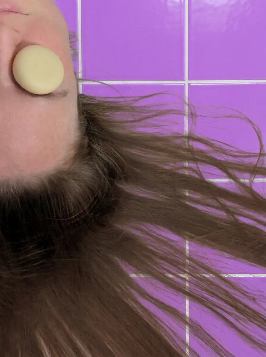 Round cream travel-size conditioner bar held over an eye, long hair on purple tiles